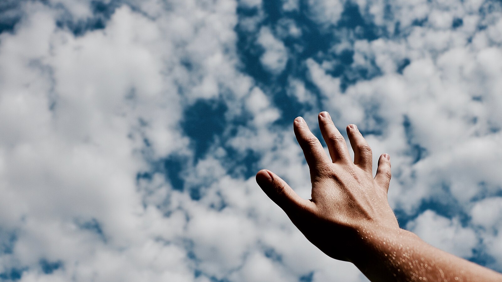 Open palmed hand reaching up towards a blue sky with puffy clouds
