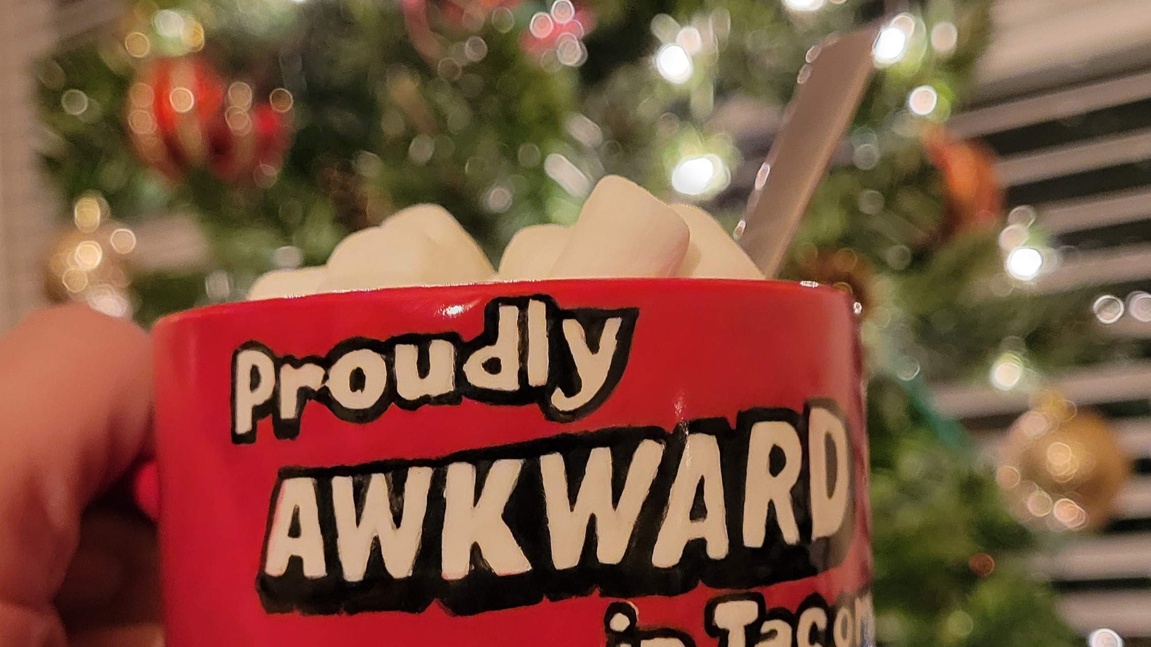 Red ceramic mug filled with hot cocoa overflowing with mini marshmallows in front of a decorated christmas tree with white lights. Mug has the text proudly awkward in white letters